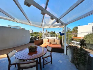 Vista de uma área externa com mesa de jantar e área de estar sob uma estrutura de pergolado.