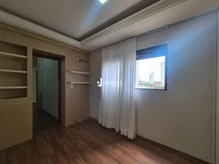 A sala apresenta piso de madeira e uma janela com cortinas brancas.