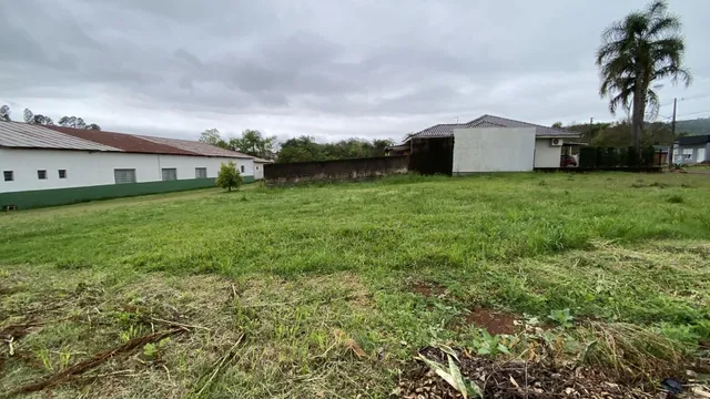 Uma foto externa mostra um terreno gramado com construções ao fundo.