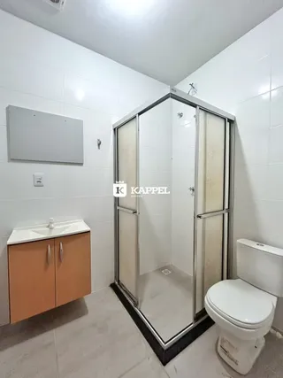 Foto interna de um banheiro com box de vidro, pias e vaso sanitário.