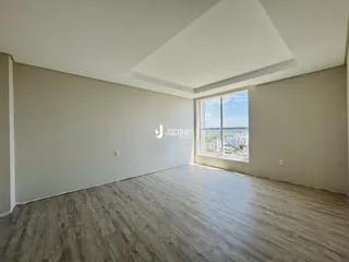 Ambiente interno de uma sala ampla com piso de madeira clara e grande janela.
