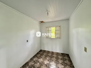 Uma foto interna mostra um cômodo com paredes brancas e piso de ladrilhos marrons.