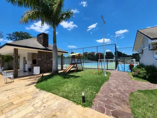 Vista externa da residência com pátio coberto, gramado e quadra esportiva no fundo.