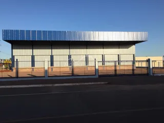 Vista externa de um galpão industrial com telhado metálico e cerca de arame.