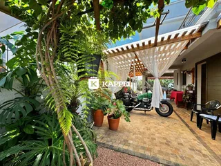 Área externa com vegetação exuberante e estrutura de pergolado sobre piso de tijolos.
