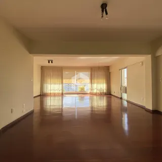 Sala ampla com piso de madeira polida e grandes janelas cobertas por cortinas.
