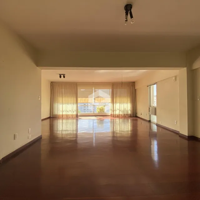 Sala ampla com piso de madeira polida e grandes janelas cobertas por cortinas.