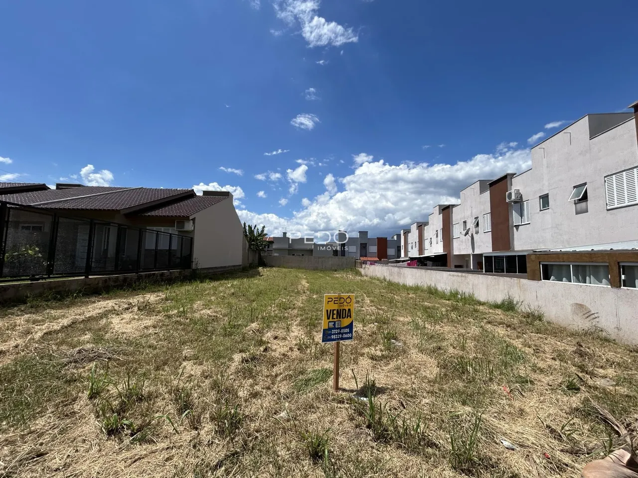 Vista externa de um terreno com grama seca e edifícios residenciais ao fundo.