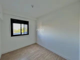 A foto mostra o interior de uma sala com piso de madeira clara e uma grande janela.