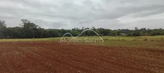 Vista de um campo aberto de terra vermelha com vegetação ao fundo.