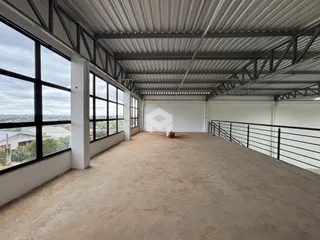 Vista interna de um grande espaço com teto de estrutura metálica e janelas amplas.