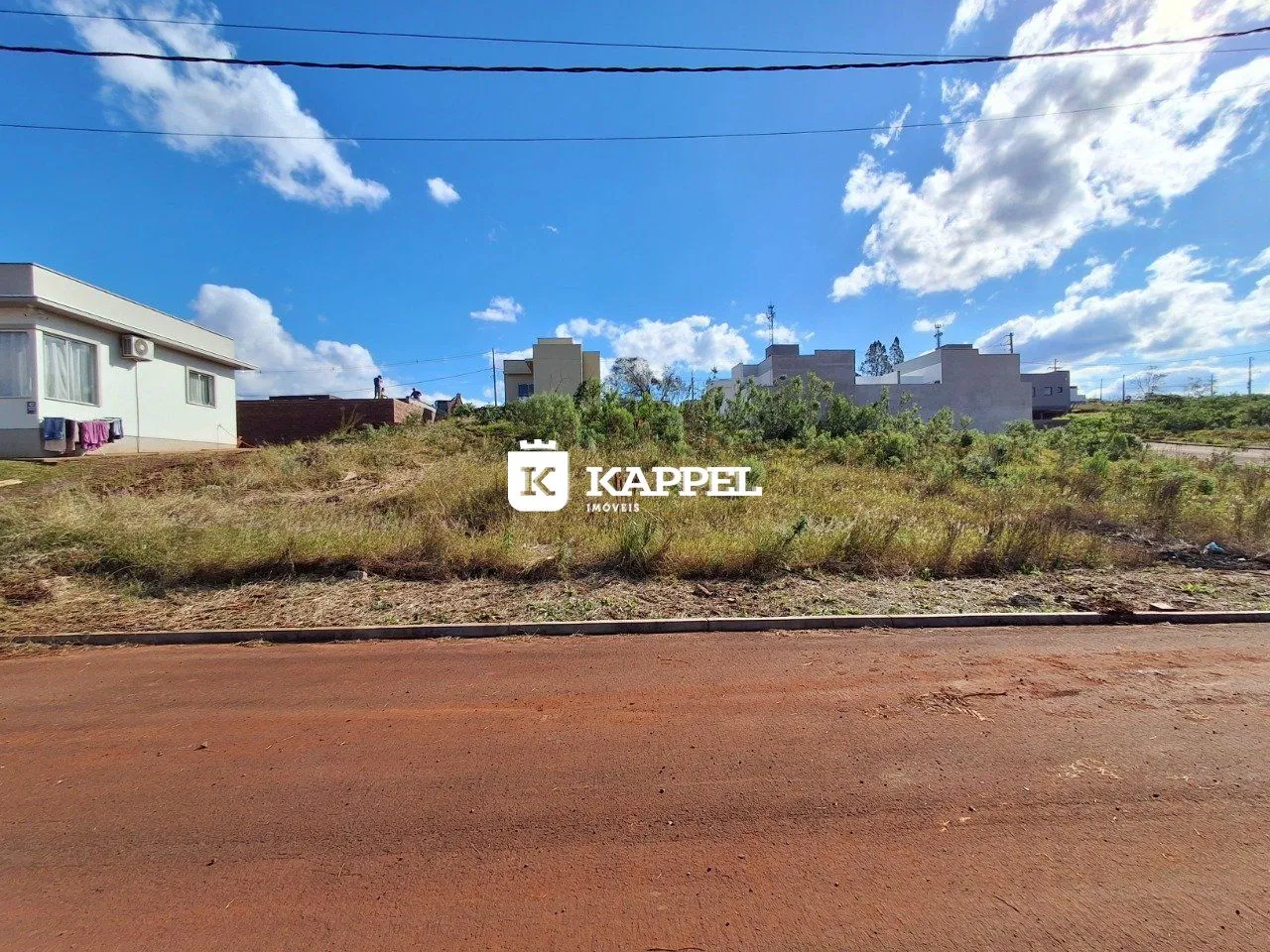 Vista externa de uma propriedade com vegetação seca e edifícios ao fundo sob um céu claro.