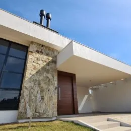 Fachada de uma residência com parede de pedra e estrutura branca sob céu azul.