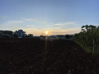 Campo de terra arada com residências ao fundo sob um céu de pôr do sol.