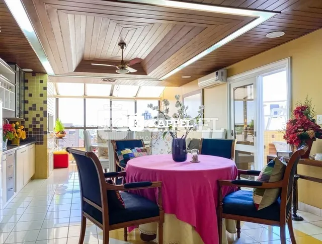 Uma mesa redonda com toalha rosa e cadeiras está posicionada em um ambiente interno com forro de madeira.