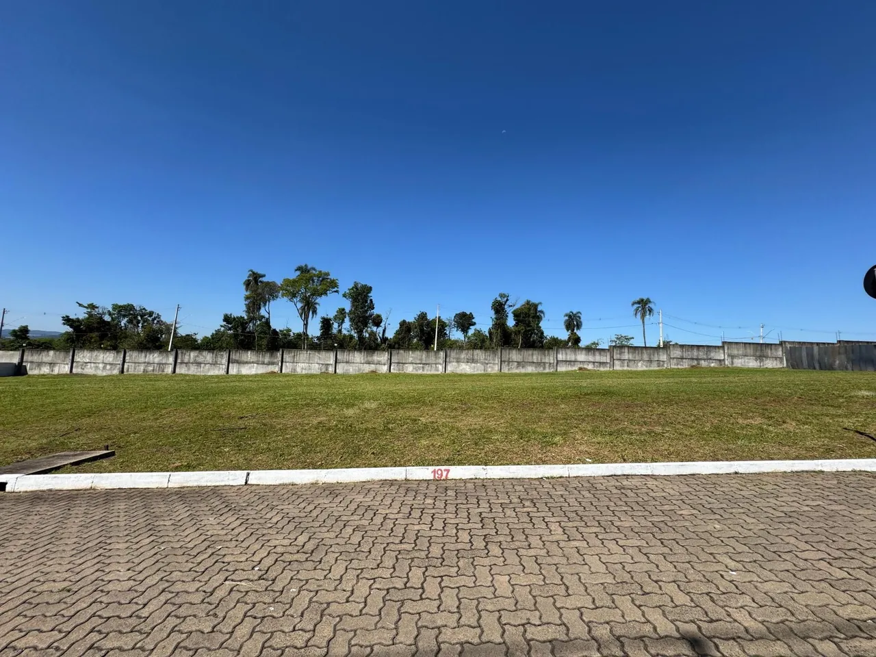 Vista externa de um campo gramado com cerca e vegetação sob um céu azul claro.