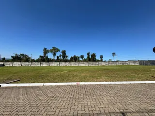 Vista externa de um campo gramado com cerca e vegetação sob um céu azul claro.
