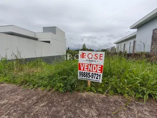 Vista externa de uma propriedade com vegetação alta e sinal de venda.