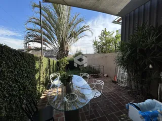 Vista de um pátio externo com mesa e cadeiras em uma área ajardinada.
