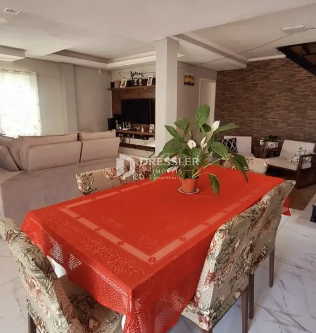 Uma mesa de jantar com toalha vermelha e cadeiras decorativas é visível no interior da sala.