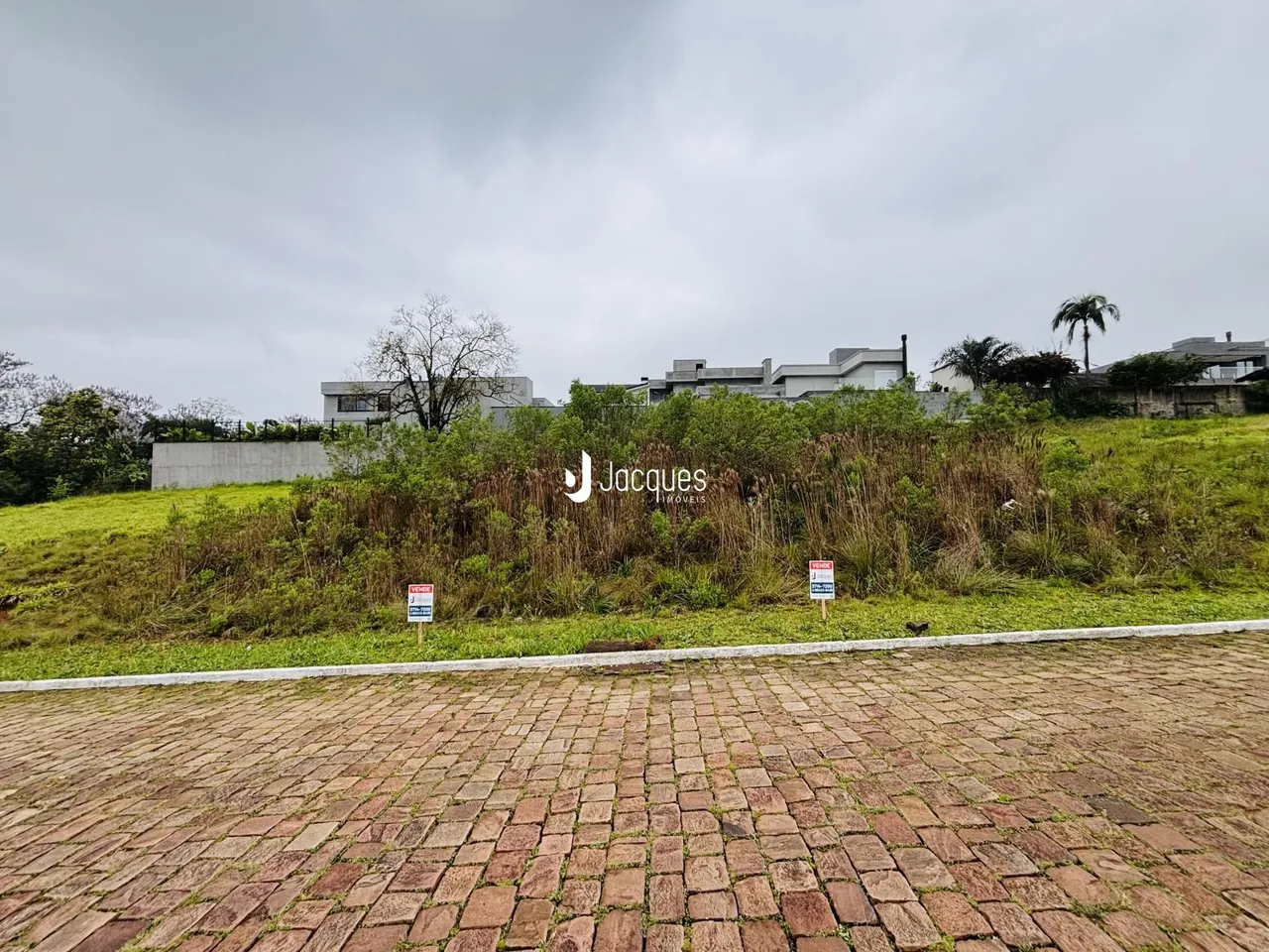Vista externa de uma propriedade com vegetação alta e piso de tijolos no primeiro plano.