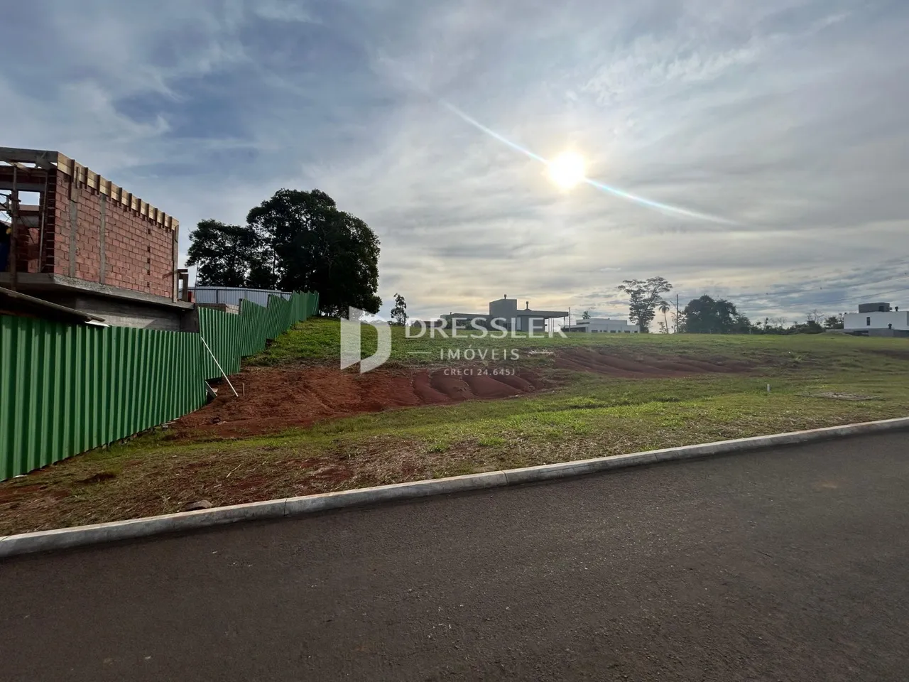 Vista externa de um terreno com cerca verde e vegetação ao lado de uma estrada.
