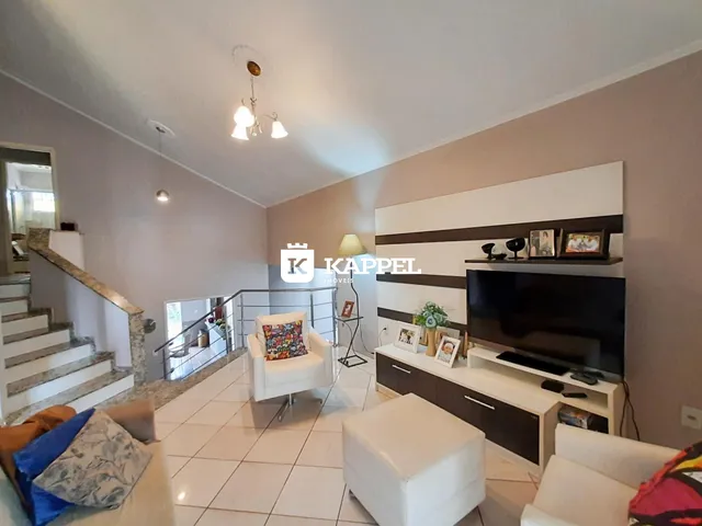 Sala de estar com sofá branco e televisão na parede, com escadaria ao fundo.