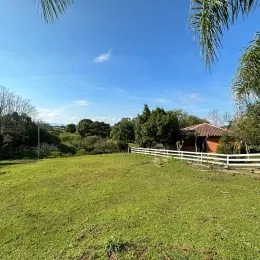 Campo gramado com cerca branca e vegetação ao fundo.