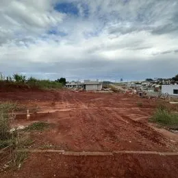 Vista de um terreno aberto com vegetação rasteira e edifícios ao fundo.