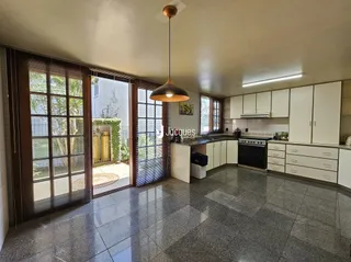 Sala de estar com janelas de madeira e cozinha ao fundo.