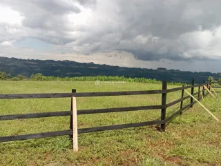 Uma cerca de madeira atravessa um campo verde com colinas ao fundo.