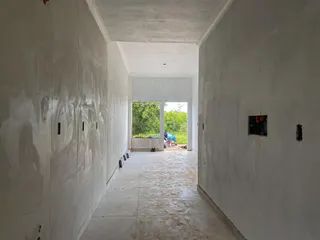 Um corredor interno com paredes brancas e piso de cerâmica leva a uma área externa verde.