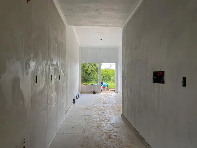 Um corredor interno com paredes brancas e piso de cerâmica leva a uma área externa verde.