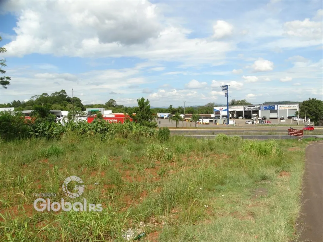Vista externa de um terreno com vegetação em primeiro plano e estruturas industriais ao fundo.