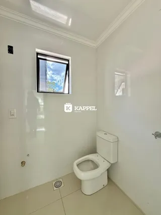 Uma foto interna mostra uma parede branca com uma janela e um vaso sanitário.