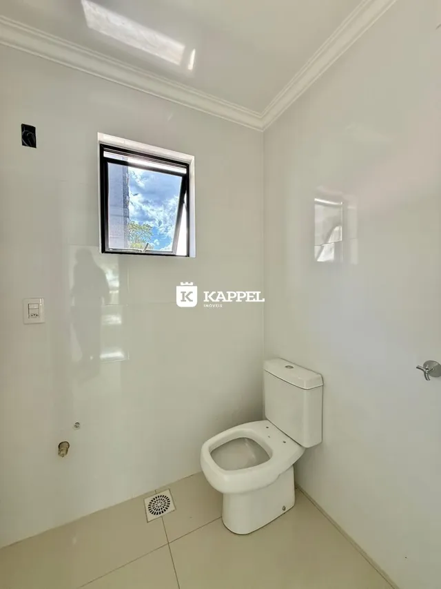Uma foto interna mostra uma parede branca com uma janela e um vaso sanitário.