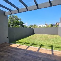 Vista de um jardim com gramado e deck de madeira sob uma estrutura de cobertura.