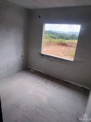 Foto interna de um cômodo com paredes brancas e uma grande janela mostrando uma paisagem externa.