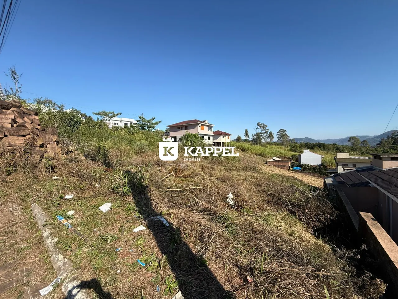 Vista externa de uma área de terreno com vegetação seca e casas ao fundo.