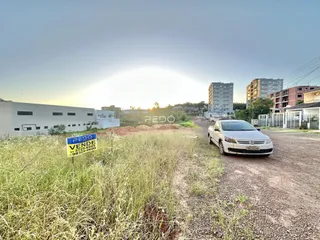 Vista externa de um terreno com vegetação alta e edifícios ao fundo.