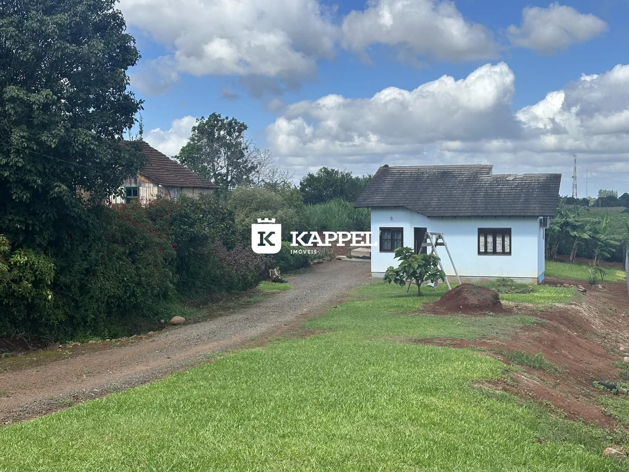 Vista externa de uma casa branca em uma área rural com gramado e estrada de terra.