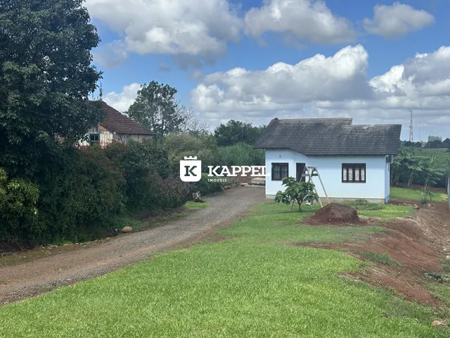 Vista externa de uma casa branca em uma área rural com gramado e estrada de terra.