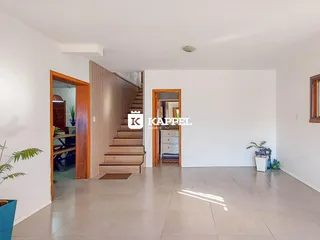 Uma sala de estar ampla com piso claro e escadaria de madeira visível.