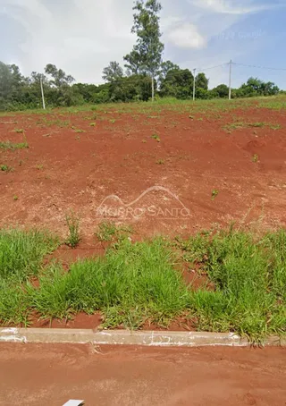 Vista de um terreno com solo avermelhado e vegetação rasteira na frente.