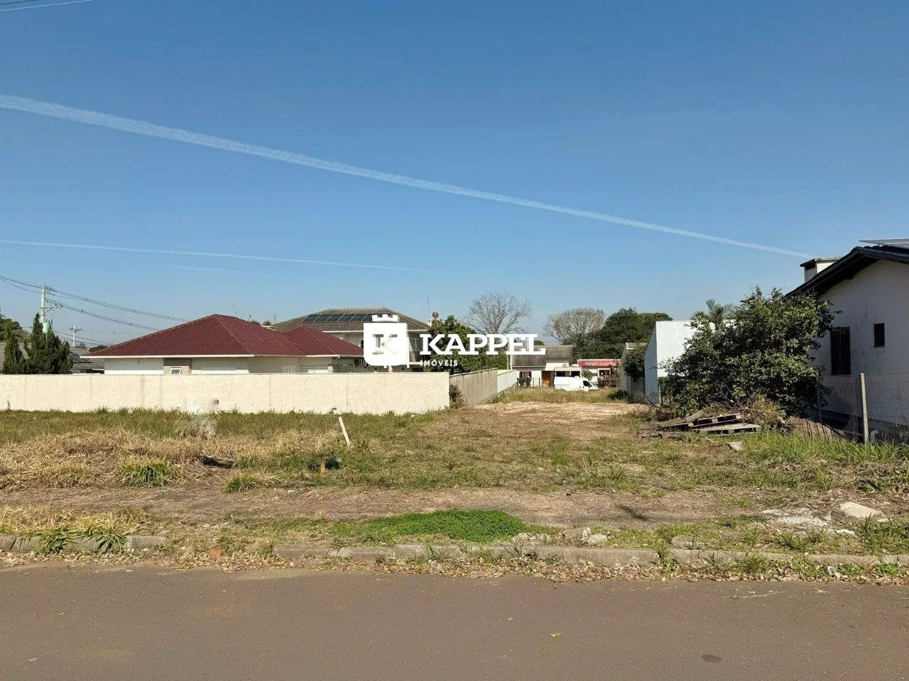 Vista externa de um terreno com vegetação seca e edifícios ao fundo.