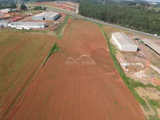 Vista aérea de uma área rural com campos de terra vermelha e estruturas industriais ao fundo.