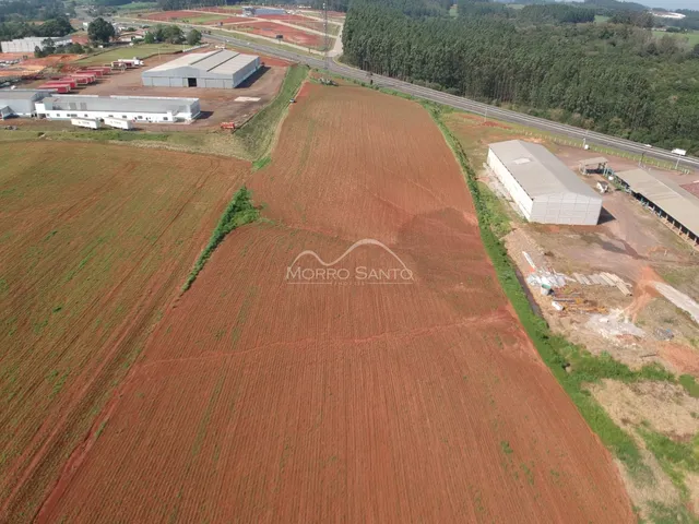 Vista aérea de uma área rural com campos de terra vermelha e estruturas industriais ao fundo.