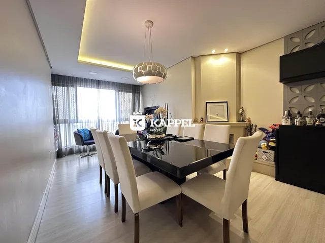 Uma sala de jantar com mesa escura e cadeiras brancas é vista em um ambiente claro.