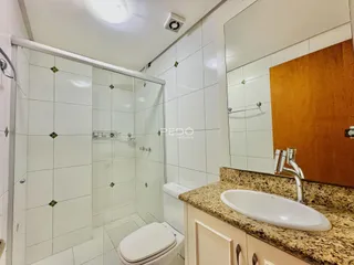 Banheiro com box de vidro, piso e paredes brancas e bancada de granito.