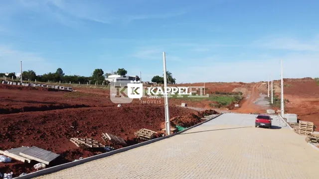 Vista de um terreno aberto com solo avermelhado e área pavimentada.